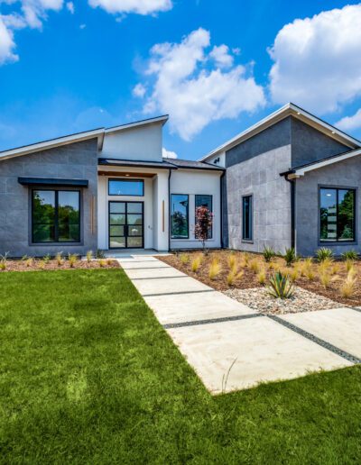 Modern single-story home with a gray exterior, large windows, and a concrete pathway leading to the entrance under a clear blue sky.
