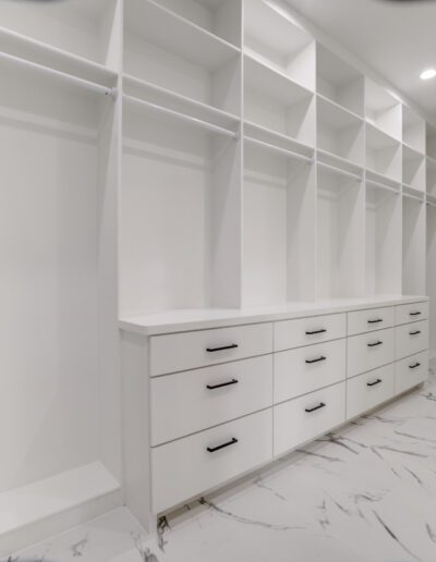 A modern, empty walk-in closet with white shelving and drawer units and marble flooring.