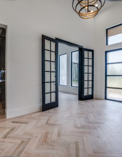 Modern interior with herringbone wood flooring, black-framed glass doors, and a view into a kitchen area.
