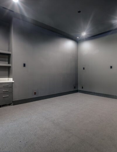 A spacious, empty room with gray walls, ceiling-mounted lights, carpet flooring, and an empty shelving unit on the left side.