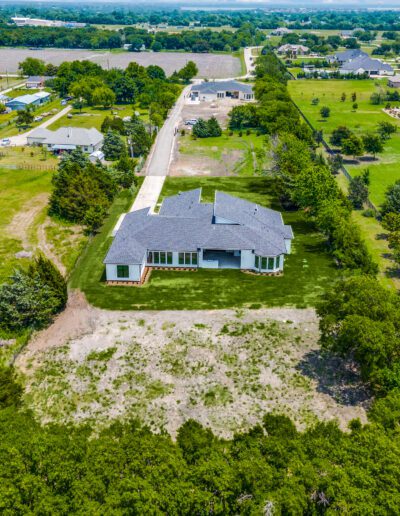 Aerial view of an isolated house with a grey roof in a large, green field, surrounded by scattered trees and neighboring homes in the distance.