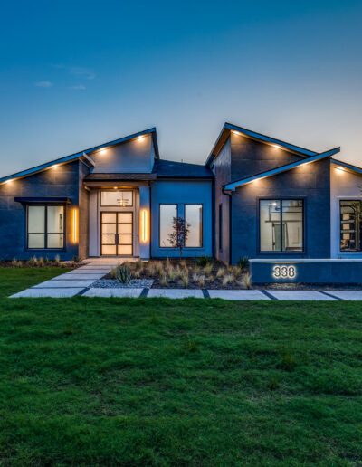 Modern single-story house with blue siding, lit up at dusk with a well-manicured lawn.