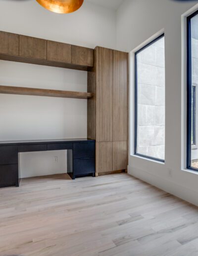 A modern home office with wooden shelves, a black chair, large windows, and beige curtains.
