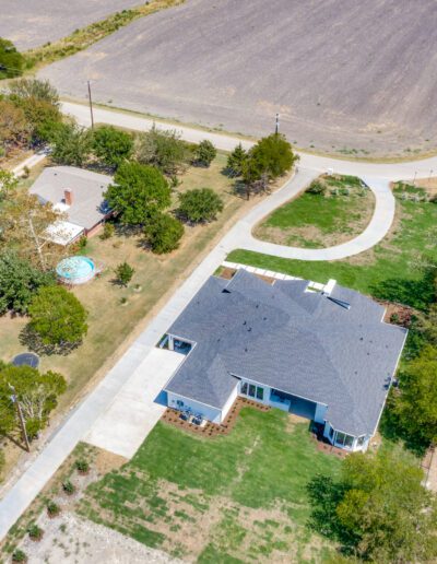 Aerial view of a suburban house with a blue roof, a curved driveway, and a backyard pool, surrounded by open land.